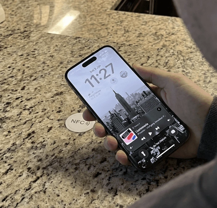 Eine Hand hält ein iPhone neben einen kleinen runden NFC-Sticker auf einer Küchenarbeitsplatte, auf dem Sperrbildschirm läuft Musik