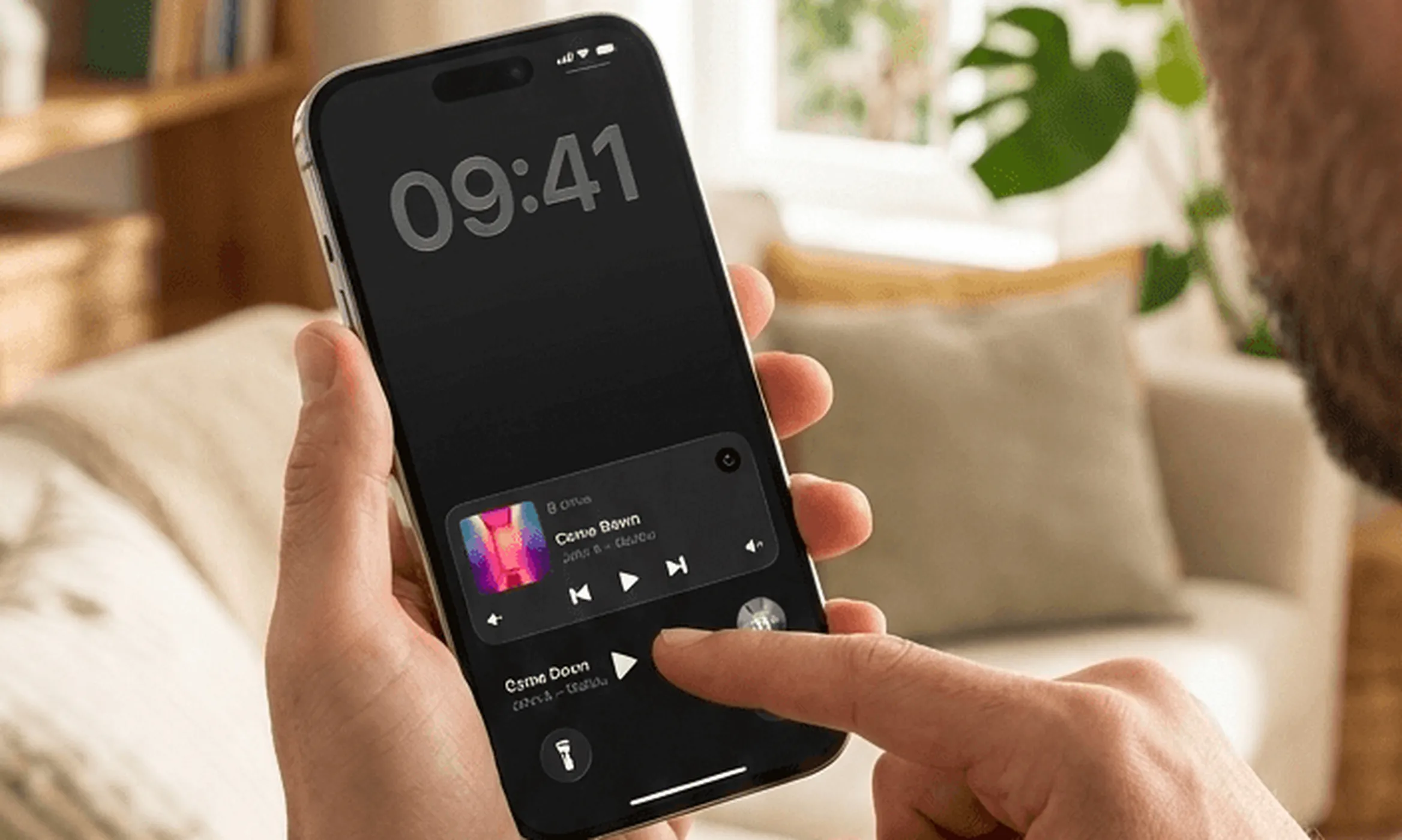 Eine Hand hält ein iPhone im Wohnzimmer, der Daumen auf einer Live-Aktivität von Lyd auf dem Sperrbildschirm mit aktuellem Titel und Wiedergabetasten